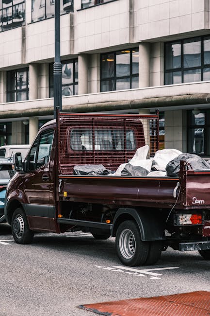 A dark red flatbed utility truck parked on a city street in front of a modern office building with large glass windows and a stone facade. The truck's open cargo area is loaded with various discarded items, including several large black plastic bags filled with waste, miscellaneous debris, and potentially recyclable materials. A metal guardrail encloses the cargo bed, providing separation from the driver’s cab, which features a black side mirror and a visible tire with a silver rim. The surroundings include other parked vehicles and urban infrastructure such as a black lamppost and painted road markings. The lighting suggests a cloudy day, with diffuse natural light illuminating the scene. This image exemplifies private waste collection or rubbish removal services, highlighting an on-site clearance process that might be part of an independent rubbish removal effort facilitated by companies like Rubbish Removal Haringey, serving areas like Alexandra Palace in Haringey.