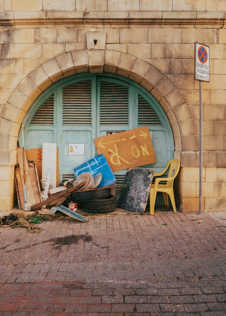 In front of a large, arched, green wooden door set within a beige brick wall, there is a collection of dumped rubbish and waste materials. This includes a yellow plastic chair positioned to the right, a blue plastic container lying on its side, a black tire, and various pieces of wood and cardboard, some leaning against the wall. A large painted yellow sign with the word 'NO' in red letters is also part of the refuse. Additionally, there are smaller items such as a paint can and a piece of black foam or mat. The waste appears haphazardly piled on the brick pavement, indicative of an unauthorized or private rubbish disposal. To the upper right of the image, a small metal pole supports a no parking or stopping sign, suggesting the area is along a street or roadway. The scene reflects a typical example of on-site rubbish accumulation and highlights the type of waste that services like Rubbish Removal Haringey often clear from private or neglected spaces, supporting alternative waste management solutions outside of standard municipal collection methods.