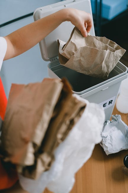 A person wearing a white shirt and red apron is disposing of a crumpled brown paper bag into a white bin with a black interior, which is placed on a wooden surface. In the foreground, there are additional crumpled paper bags, some partially folded and others untidily bunched together. The bin is situated outdoors or in a well-lit area, with a background showing a dark, blurred surface, possibly pavement or a pathway. The scene depicts a typical household or commercial waste disposal activity, fitting within the context of rubbish removal or on-site waste management services provided by Rubbish Removal Haringey, illustrating the manual process of gathering and discarding recyclable or general rubbish using paper packaging materials, set against a clean and organised environment.