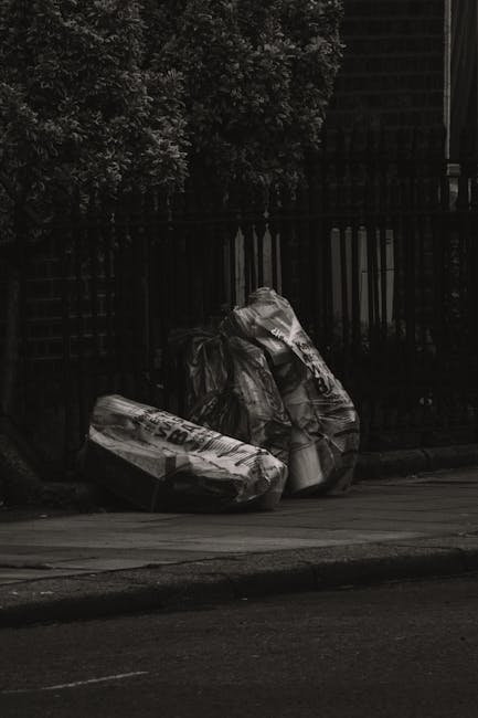 Two large black plastic rubbish bags, slightly crumpled and tied at the top, are leaning against a black metal fence on a pavement in an outdoor urban setting. The bags appear to contain household waste or debris, with some items protruding slightly from the open tops. Behind the fence, there are dense green shrubs or trees with textured foliage, and a dark brick wall or building is partially visible in the background. The scene is lit with natural daylight, casting soft shadows on the ground, indicating a typical street environment suitable for waste collection or clearance services. The presence of these bags suggests private or independent waste disposal, in contrast to municipal rubbish collection, supporting services like those offered by Rubbish Removal Haringey in the local area.