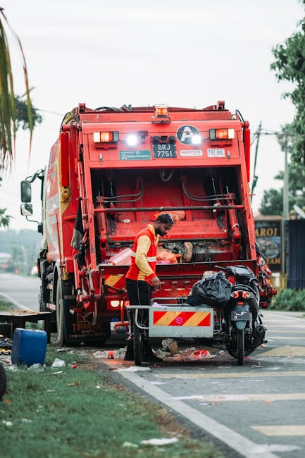 A waste collection worker in a red and yellow safety uniform is standing next to a large red rubbish truck on a roadside. The back of the truck, which features a mechanical compactor, is open, revealing the interior where waste is being loaded or managed. The worker appears to be handling or inspecting waste materials, which are not clearly visible but include a black plastic bag resting on the side of the vehicle. The scene is set on a paved street, with a small section of grassy verge and sidewalk visible in the foreground. In the background, there are trees, utility poles, and signage indicating a commercial or urban area. The lighting suggests daylight hours with overcast skies, and the overall scene depicts an effort related to on-site waste collection, highlighting the importance of efficient rubbish removal services that could be provided by companies like Rubbish Removal Haringey.