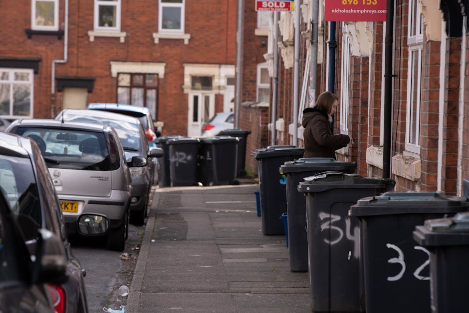 Two large black plastic rubbish bags, slightly crumpled and tied at the top, are leaning against a black metal fence on a pavement in an outdoor urban setting. The bags appear to contain household waste or debris, with some items protruding slightly from the open tops. Behind the fence, there are dense green shrubs or trees with textured foliage, and a dark brick wall or building is partially visible in the background. The scene is lit with natural daylight, casting soft shadows on the ground, indicating a typical street environment suitable for waste collection or clearance services. The presence of these bags suggests private or independent waste disposal, in contrast to municipal rubbish collection, supporting services like those offered by Rubbish Removal Haringey in the local area.