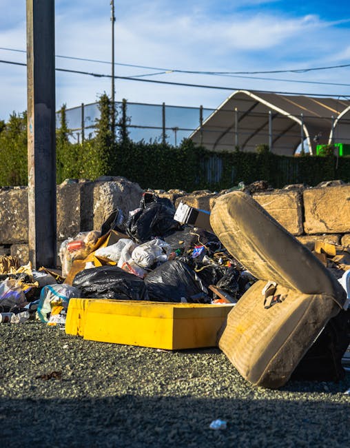 A pile of mixed waste including black garbage bags, a yellow plastic bin, and an old, worn car tire lying on a gravel surface in an outdoor area. The waste is situated near a low stone wall with a weathered wooden post visible on the left side of the image. Behind the waste, there is a dark green hedge, which partially obscures a metal fence and a large, white, semi-transparent canopy or tent structure. Overhead, power lines stretch across a partly cloudy sky, indicating an urban or suburban environment. The scene suggests an improper disposal of rubbish, highlighting a need for professional rubbish removal services such as those offered by Rubbish Removal Haringey, which can facilitate alternative waste handling and on-site clearance, preventing accumulation of waste in communal or private spaces.