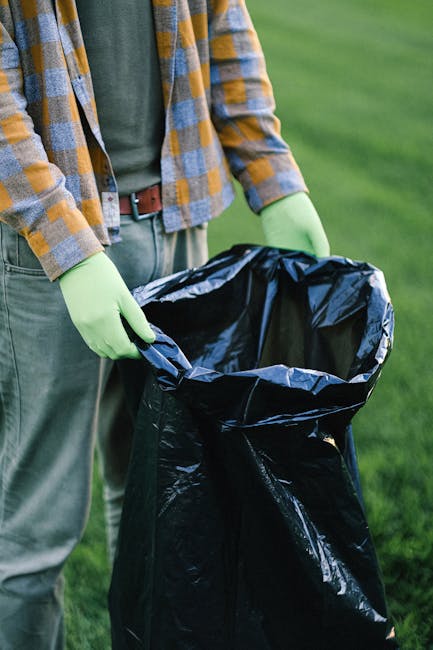 A person standing outdoors on a grassy area is holding open a large black rubbish bag made of thick plastic material, with the bag partially filled and collapsing slightly inward. The individual is wearing a brown and yellow checkered shirt over a green top, blue jeans, and bright yellow-green gloves designed for waste handling. The background features a well-maintained lawn or park space with a soft, natural green tone. The scene suggests an activity related to waste collection or clearance, possibly associated with private or independent rubbish removal services, such as those provided by Rubbish Removal Haringey, which specializes in responsible waste disposal. The setting is bright with natural daylight, emphasizing clarity and cleanliness in the process of rubbish collection or disposal, aligned with non-authority waste management options common in private rubbish removal and on-site clearance activities.