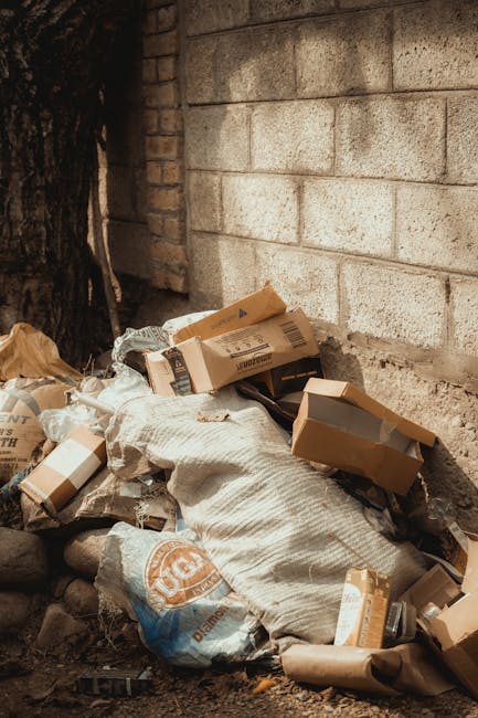 A pile of mixed waste including black garbage bags, a yellow plastic bin, and an old, worn car tire lying on a gravel surface in an outdoor area. The waste is situated near a low stone wall with a weathered wooden post visible on the left side of the image. Behind the waste, there is a dark green hedge, which partially obscures a metal fence and a large, white, semi-transparent canopy or tent structure. Overhead, power lines stretch across a partly cloudy sky, indicating an urban or suburban environment. The scene suggests an improper disposal of rubbish, highlighting a need for professional rubbish removal services such as those offered by Rubbish Removal Haringey, which can facilitate alternative waste handling and on-site clearance, preventing accumulation of waste in communal or private spaces.