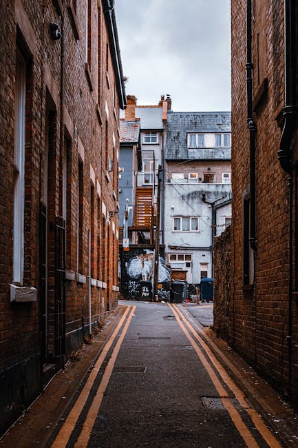 A narrow urban alleyway situated between two tall brick buildings with reddish-brown facades, featuring visible drainpipes attached along the walls. The asphalt surface of the alley is marked by double yellow lines running parallel each side, indicating parking restrictions. In the distant background, there is a small assortment of residential structures with varied construction materials and finishes, including grey concrete and slate roofing, some with small windows and external staircases. The sky overhead appears overcast with a muted, greyish tone, casting diffuse natural light across the scene. The alley is clean and free of debris, with no visible rubbish or waste bins present, but the overall setting suggests it is an area where private waste removal services like Rubbish Removal Haringey might operate for on-site clearance or alternative waste handling solutions in the vicinity of residential properties.