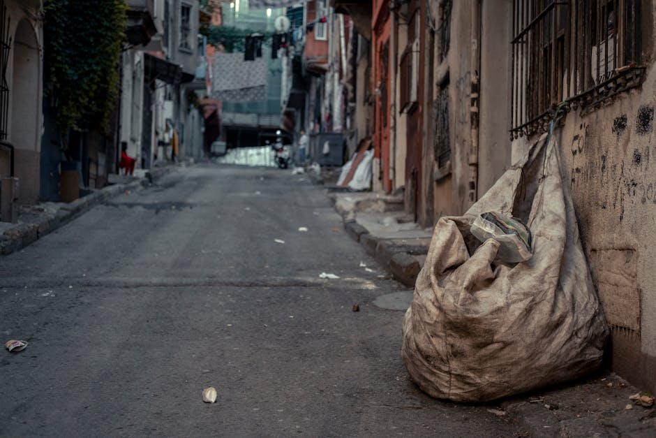 A large, weathered, beige fabric rubbish bag is positioned on the right side of a narrow, uneven urban street pavement, leaning against a graffitied wall with metal bars on the windows. The bag appears slightly overfilled, with some plastic packaging visible protruding from the top. Surrounding the bag, small pieces of litter and debris spread across the concrete surface, which looks worn and rough in texture. The background features tightly packed residential buildings with various facades, some with visible balconies or shutters, and laundry hanging on lines above the street. The scene is evenly lit, suggesting daytime with diffuse natural light, and there are no people present. This setting reflects an alternative approach to rubbish removal in a densely built area, where informal disposal methods are visible, aligning with a focus on independent waste handling around urban environments.