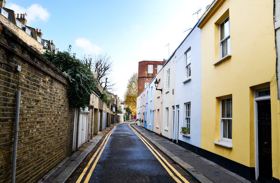 A narrow urban alleyway situated between two tall brick buildings with reddish-brown facades, featuring visible drainpipes attached along the walls. The asphalt surface of the alley is marked by double yellow lines running parallel each side, indicating parking restrictions. In the distant background, there is a small assortment of residential structures with varied construction materials and finishes, including grey concrete and slate roofing, some with small windows and external staircases. The sky overhead appears overcast with a muted, greyish tone, casting diffuse natural light across the scene. The alley is clean and free of debris, with no visible rubbish or waste bins present, but the overall setting suggests it is an area where private waste removal services like Rubbish Removal Haringey might operate for on-site clearance or alternative waste handling solutions in the vicinity of residential properties.