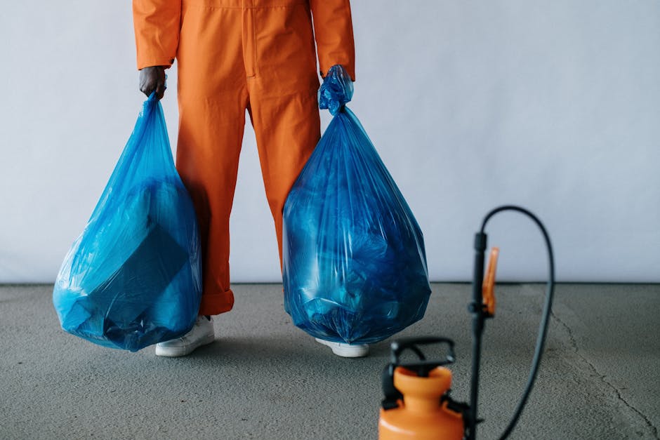 A person dressed in an orange waste collection uniform stands on a concrete floor indoors, holding two large blue plastic rubbish bags, one in each hand. The bags are filled with waste and are slightly translucent, showing the varying shapes and sizes of contents inside. The individual is wearing white footwear and grey gloves, with their lower legs visible, but their face and upper body are not shown. In the foreground, a small professional-grade spray or spray gun with an orange handle and black hose is partially visible, resting on the floor. The background features a plain light grey wall, creating a neutral setting typical for waste collection or clearance services. The scene reflects the process of on-site rubbish removal, consistent with private waste management activities such as those provided by Rubbish Removal Haringey, and illustrates the physical handling of waste materials during a clearance operation, supporting the context of alternative disposal methods outside local authority collection.