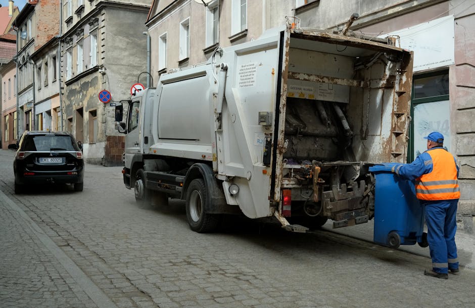 A worker wearing a blue uniform and a high-visibility orange vest is emptying a blue recycling bin into the rear compartment of a white waste collection truck parked on a narrow urban street. The truck's rear loading area is open, revealing a rusty, metal interior designed for compacting refuse, with visible mechanical components and controls. To the left of the truck, a black car is parked close to the curb. The surrounding environment includes old, multi-story buildings with weathered facades, some showing signs of peeling paint and age-related wear. The street is paved with cobblestones, and there are no other vehicles or pedestrians visible. The overall scene depicts a routine rubbish collection operation, representing private waste removal activity typical of alternative disposal methods outside of municipal services, as occasionally undertaken by companies like Rubbish Removal Haringey. The lighting suggests a daytime setting with natural ambient light highlighting the textures of the materials and environment.