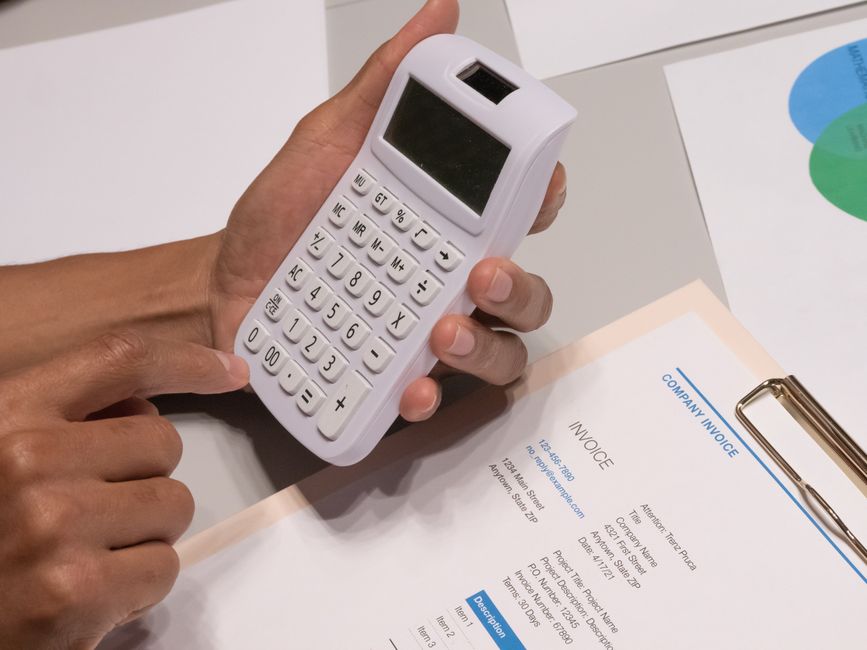 A person holding a white calculator with a large digital display, several function buttons, and numerical keys, positioned over a white table. The individual's hand is visible, with dark skin and neatly trimmed nails, pressing the calculator's buttons. To the right, there is a beige clipboard holding a printed invoice with a blue border, text, and a signature area, partially visible on the table. In the background, the edge of a white wall and part of a sheet of paper with colored charts can be seen. The scene appears to be an office environment related to financial calculations, possibly for budgeting or cost estimation in waste management or rubbish removal services, as suggested by the context of the website Rubbish Removal Haringey.