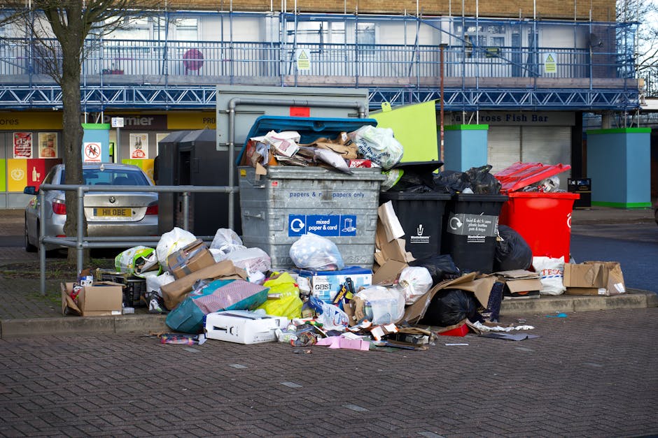 In this outdoor urban scene, a large collection of mixed household waste and recyclables has been accumulated in front of a municipal waste container on a paved parking area. The waste includes numerous black and black plastic bin bags, some toppled or partially open, revealing crumpled paper, cardboard boxes, and plastic packaging inside. Adjacent to these, there are several open-top containers; one is a grey mixed paper and cardboard bin, filled to the brim with paper slips and flattened boxes, with some waste spilling over the edges onto the ground. A black bin with white labels appears to be designated for general waste, while a bright red recycling bin nearby likely serves for recyclable materials. Surrounding the bins, discarded packaging, flattened boxes, and loose paper spread across the pavement, indicating an overflowing arrangement not suitable for regular council collection. The background features a storefront with shuttered windows and a blue construction safety barrier on the upper level, while a parked silver car is situated to the left behind a simple metal barrier, emphasizing the urban environment. This scene visually illustrates the need for efficient rubbish removal and private waste handling services, such as those provided by Rubbish Removal Haringey, to manage excess waste outside typical collection schedules.