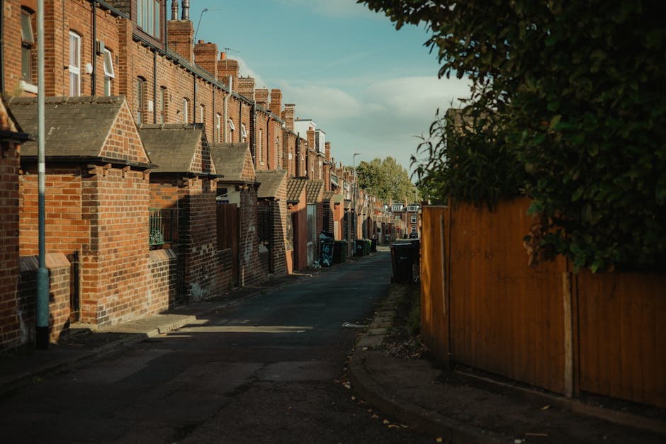 A narrow residential alleyway in a suburban area during daylight, lined with a series of brick terraced houses on the left side, featuring brick facades, pitched tiled roofs, and small front steps. The houses have small window openings and chimneys visible on rooftops. On the right side, a tall wooden fence runs alongside the alley, partially obscuring a lush green hedge that extends above it. The pavement and asphalt road are worn, with some scattered debris visible along the edges. Several wheelie bins and trash containers are placed along the front of the houses, indicating rubbish collection points. Shadows cast by the buildings and foliage create a patchwork of light and shade across the scene. The environment suggests a quiet, well-maintained neighborhood, where private waste collection — including the use of wheelie bins — is a common alternative to municipal rubbish removal, as exemplified by services like Rubbish Removal Haringey, which provides professional rubbish disposal in the area.