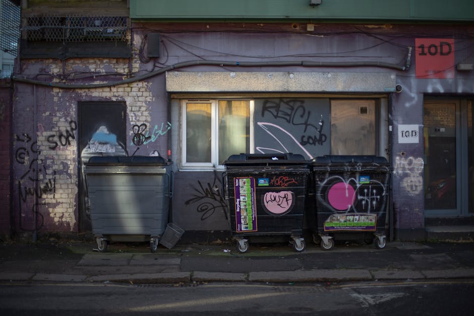 The image depicts a section of the exterior of a brick building with weathered, partially exposed bricks and graffiti in multiple colors, including black, white, pink, and blue. In the foreground, a concrete sidewalk runs parallel to the building, with signs of wear and small scattered debris. Two large wheelie bins are positioned close to the building's wall: one grey with a closed lid on the left, and one black with a graphic sticker and graffiti on the right, both fitted with plastic wheels. Behind the bins, there is a small, rectangular window with a white frame, partially covered by a beige roller shutter that is partially rolled down, obscuring part of the view inside. Above, exposed wiring and cables run horizontally across the building's façade, above a narrow ledge. To the right, part of a glass door with a grey frame and a small red and white sign marked '10D' is visible, with additional graffiti alongside it. The overall scene has a gritty urban atmosphere, illustrating an area where rubbish collection might occur as part of a private waste management service, with Rubbish Removal Haringey potentially involved in the clearing of such accumulated refuse in an alternative waste handling context.